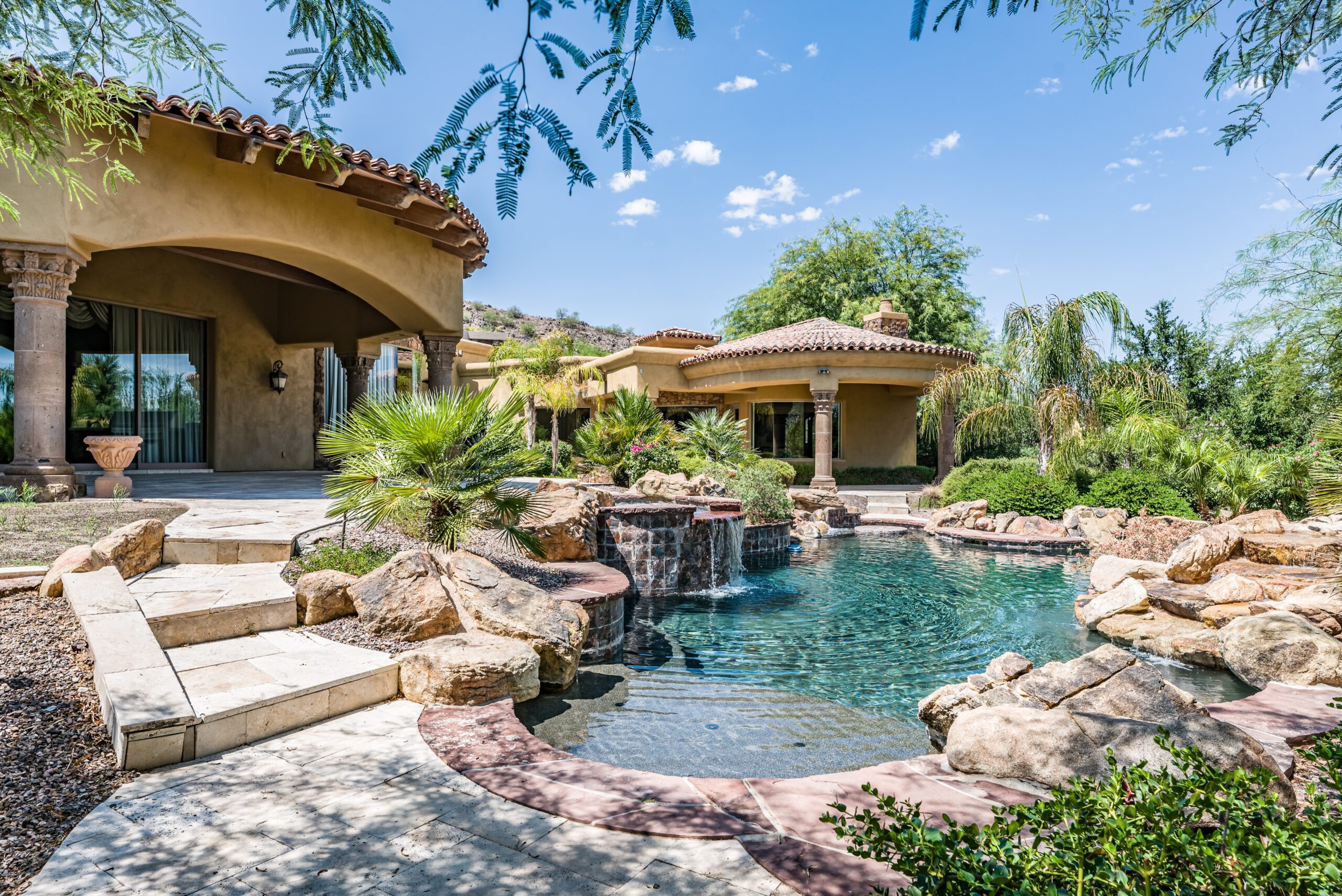 A patio with a dining table and chairs on stone flooring, adjacent to a house with French doors. It is surrounded by lush greenery and a vibrant flower arrangement in a large pot.